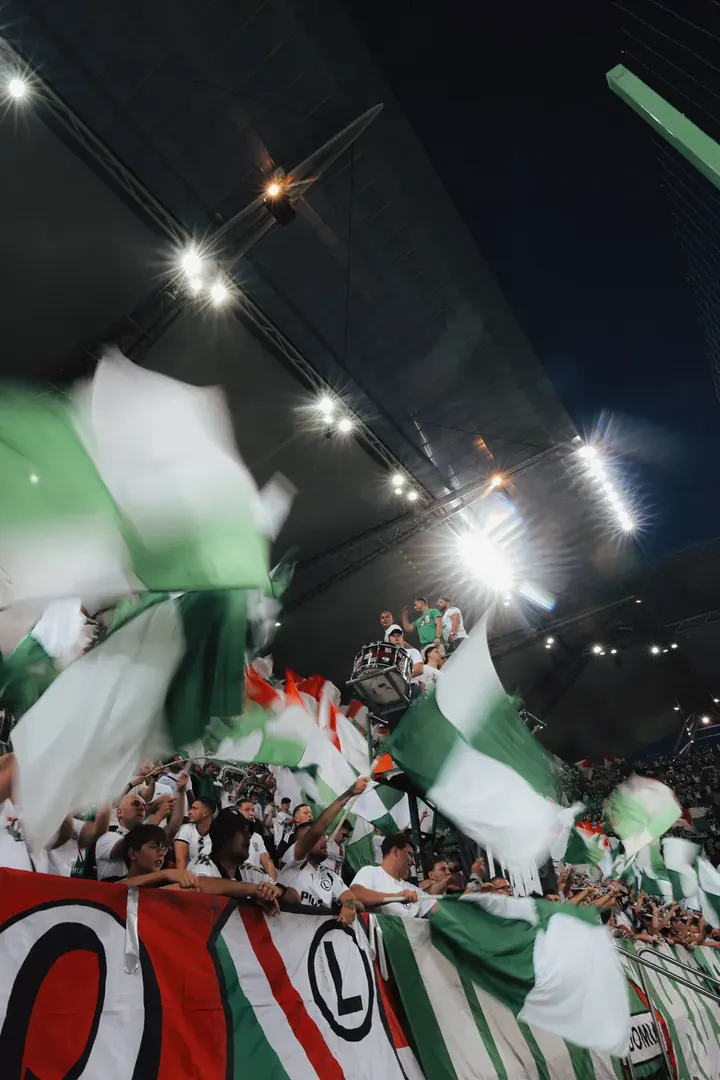 Kibice machają zielono-białymi flagami pod dachem stadionu